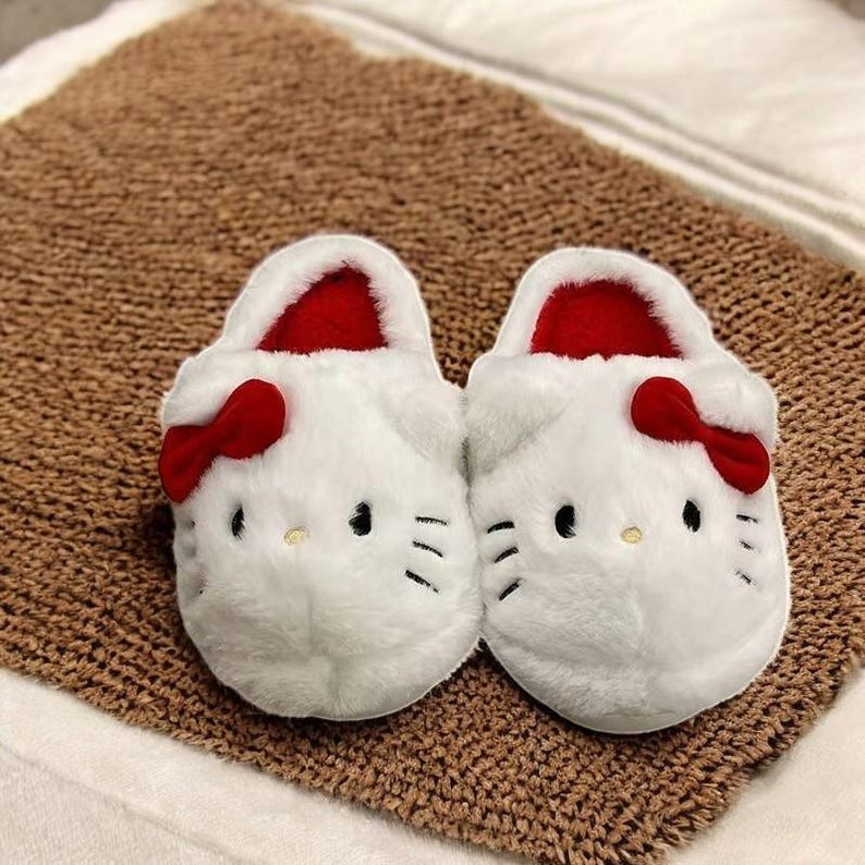 May include: A pair of white fuzzy slippers with red bows and a Hello Kitty face design.