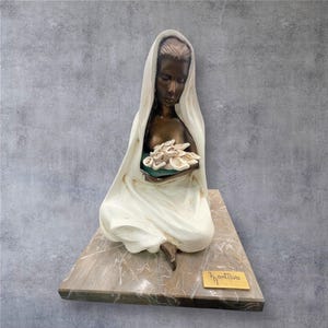 May include: A bronze sculpture of a woman sitting with a white cloth draped over her. She is holding a bouquet of white flowers in her hands. The sculpture is on a marble base with the artist's signature "Fontiva" inscribed on a gold plaque.