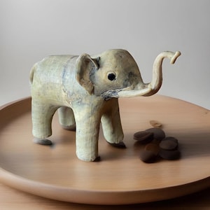 May include: A light-coloured elephant figurine with a curled trunk and large ears. The elephant is standing on a wooden tray, with a small pile of brown stones nearby. The figurine has a textured surface.