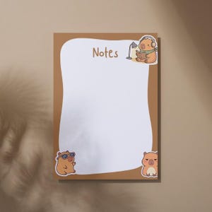May include: A brown notepad with a white interior and the word "Notes" in brown. The notepad features cartoon illustrations of a capybara wearing headphones, sunglasses, and with a grumpy expression.