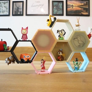 Peut inclure: Étagère en nid d'abeille géométrique moderne, noire, rose et bleue, remplie de figurines et de jouets colorés. Les étagères sont disposées sur une table en bois, avec des œuvres d'art encadrées en arrière-plan.