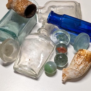 Pode incluir: Uma coleção de garrafas de vidro antigas, bolas de gude e um cachimbo de cerâmica. As garrafas variam em forma e cor, incluindo transparente, azul e verde. Várias bolas de gude são visíveis, com redemoinhos vermelhos e verdes. O cachimbo de cerâmica é bege e branco.