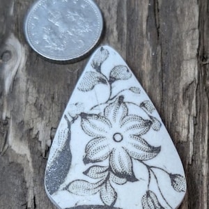 May include: A white ceramic tile with a black and white floral design. The tile is shaped like a teardrop and has a smooth, glossy finish.