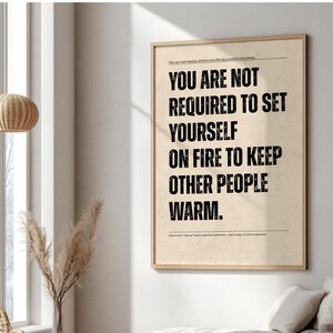 May include: A framed print with the text "YOU ARE NOT REQUIRED TO SET YOURSELF ON FIRE TO KEEP OTHER PEOPLE WARM." The text is in black, centered on a beige background, and set in a simple sans-serif font. The frame is a light brown colour.