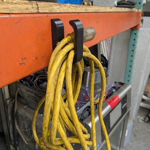 Op de afbeelding: Een zwarte, 3D-geprinte koordhouder bevestigd aan een oranje metalen plank, met een opgerold geel verlengsnoer. Het snoer heeft een standaard driepolige stekker. De plank heeft een houten bovenkant en bevindt zich in een werkplaats.