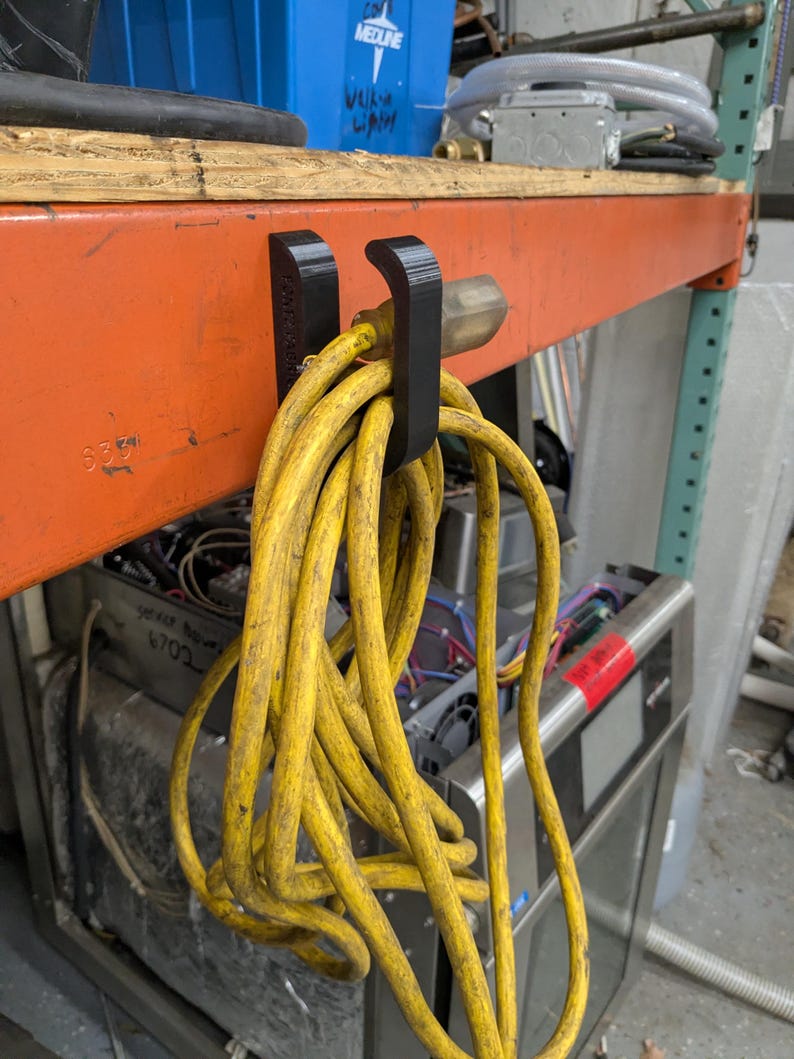Op de afbeelding: Een zwarte, 3D-geprinte koordhouder bevestigd aan een oranje metalen plank, met een opgerold geel verlengsnoer. Het snoer heeft een standaard driepolige stekker. De plank heeft een houten bovenkant en bevindt zich in een werkplaats.