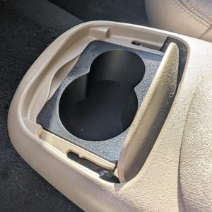 May include: A tan car console with a black insert designed to hold cups. The insert has two cup holders and is labeled "Removable". The console lid is partially open, revealing the cup holders.