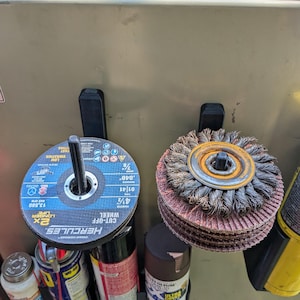 Puede incluir: Una colección de herramientas y suministros, que incluye una rueda de corte Hercules, una rueda de alambre y varias latas de pintura en aerosol y lubricantes. Los artículos están organizados en soportes negros fijados a una superficie metálica.