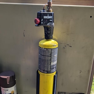 May include: A black and yellow Bernzomatic TS4000 torch head attached to a yellow propane cylinder. The torch has a red control knob and is mounted on a metal surface. A can of spray paint is visible to the left.