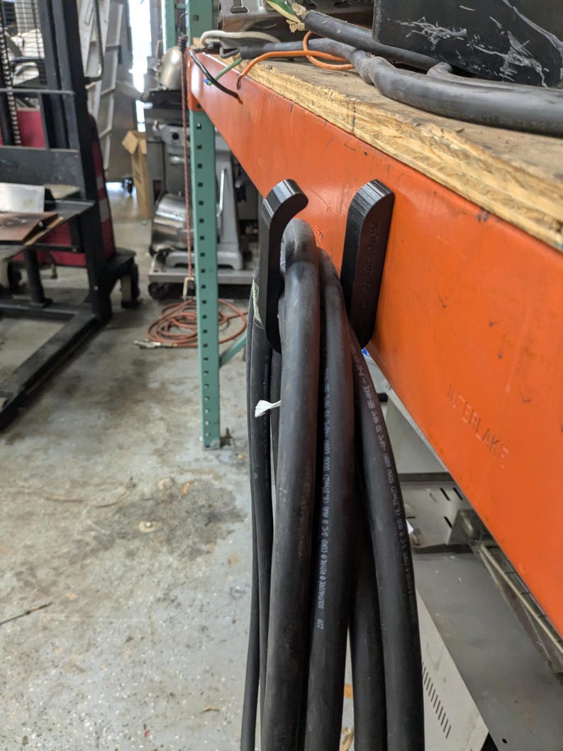 Op de afbeelding: Zwarte elektrische kabels hangen aan zwarte haken die aan een oranje plank zijn bevestigd. De plank is gemaakt van hout en metaal. De kabels zijn dik en hebben witte tekst erop gedrukt. De achtergrond is een werkplaats.