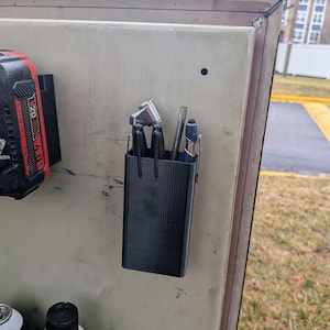 May include: A black, 3D-printed tool holder with multiple compartments, mounted on a beige metal surface. The holder contains various tools, including a wrench and a pen. A red and black battery pack is also visible.
