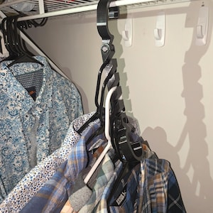 May include: A black multi-hanger system is attached to a white wire shelf, holding several shirts. The shirts are various colors and patterns, including blue floral and plaid designs. The hanger system appears to be made of plastic.