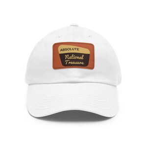 Puede incluir: Gorra de béisbol blanca con un parche de cuero marrón que dice "ABSOLUTE NATIONAL TREASURE" en texto amarillo.