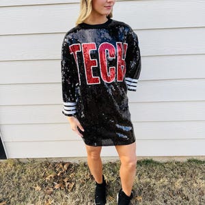 May include: A black sequin mini dress with a red and white "TECH" graphic. The dress has a short sleeve and a round neckline.