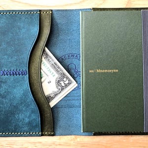 May include: A bi-fold leather wallet in blue and dark green featuring a pocket with blue stitching, a slot containing a two-dollar bill, and a dark green Mnemosyne notebook with a vertical elastic band and gold text on the front cover.