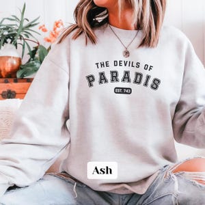 May include: A light gray sweatshirt with the text "THE DEVILS OF PARADIS EST. 743" printed on the front. The sweatshirt is worn by a person with long brown hair. The word "Ash" is printed on the bottom of the sweatshirt.