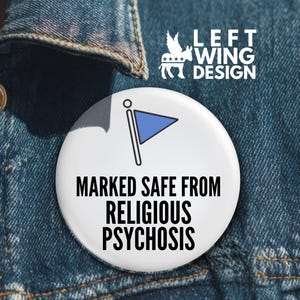 Puede incluir: Una insignia blanca con el texto "MARKED SAFE FROM RELIGIOUS PSYCHOSIS" y un gráfico de bandera azul. La insignia está prendida a una chaqueta vaquera. La esquina superior derecha tiene el texto "LEFT WING DESIGN" con un gráfico.