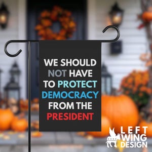 May include: A black rectangular garden flag hanging on a metal stand. The text reads: WE SHOULD NOT HAVE TO PROTECT DEMOCRACY FROM THE PRESIDENT. The words are in white, light blue, and red, with the brand name LEFT WING DESIGN in the corner.