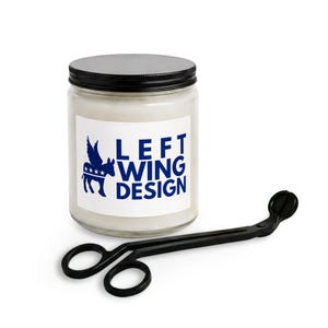 May include: A clear glass jar candle with a black lid and a white label featuring a blue graphic of a winged donkey with four stars and the text LEFT WING DESIGN in blue. A black metal wick trimmer is positioned in front of the candle.