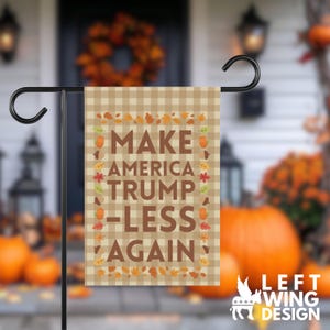 May include: A rectangular garden flag featuring a beige and tan gingham pattern background. The text "MAKE AMERICA TRUMP-LESS AGAIN" is printed in large brown capital letters. A border of colorful autumn leaves, pumpkins, and acorns surrounds the text.