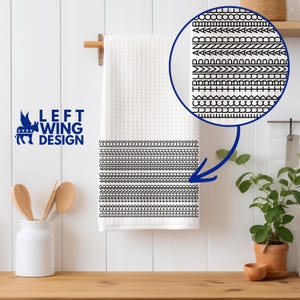 May include: White waffle-weave towel with a black geometric border design. The towel is hanging on a wooden rod. The text "LEFT WING DESIGN" is printed in blue on the left side of the towel.