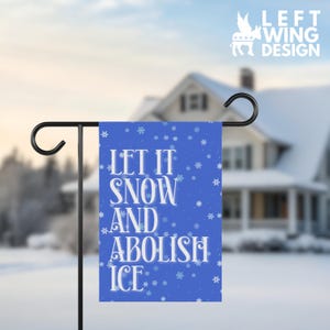 May include: A blue garden flag with white text that reads "LET IT SNOW AND ABOLISH ICE." The flag has a snowflake pattern and is displayed on a black metal stand. The upper right corner has the text "LEFT WING DESIGN" with a donkey logo.