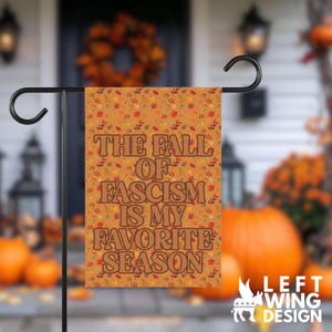 May include: A rectangular garden flag featuring an orange background with a repeating pattern of autumn leaves and berries. The text THE FALL OF FASCISM IS MY FAVORITE SEASON is printed in large, bold, brown, retro-style lettering across the centre.