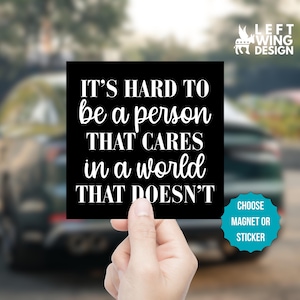 May include: A black square magnet or sticker with white text that reads "IT'S HARD TO be a person THAT CARES in a world THAT DOESN'T." The design includes a logo and the option to choose a magnet or sticker.