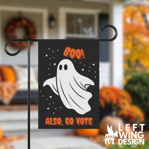 May include: A rectangular black garden flag featuring a white ghost illustration in the center. The text BOO! appears in orange above the ghost, and the text ALSO, GO VOTE appears in orange below the ghost. Small white dots are scattered around.