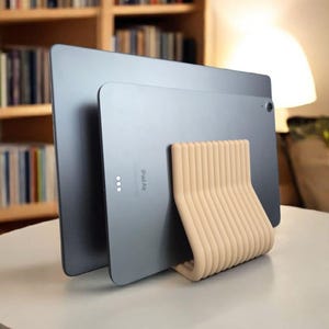 May include: Two gray iPad Air tablets are propped up by a beige, curved stand on a white table. The stand has a series of vertical, rounded supports. The tablets are positioned in front of a bookshelf and a lamp.