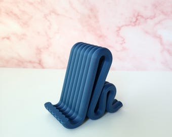 May include: A matte navy blue phone stand featuring a continuous, serpentine, S-shaped folded design. The structure consists of multiple parallel, rounded ridges that form a stable base and an angled support ledge for holding a mobile device.