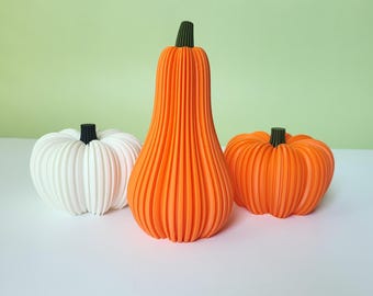 May include: Three decorative pumpkins in a row. The pumpkin on the left is white with a black stem. The tall center pumpkin is orange with a green stem. The pumpkin on the right is orange with a green stem. All three pumpkins have vertical ridges.