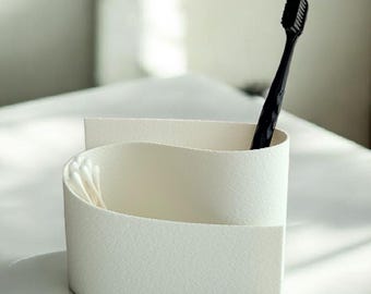 May include: A white, textured, minimalist toothbrush holder with a curved design. A black toothbrush and cotton swabs are visible inside. The holder sits on a white surface, with soft lighting in the background.