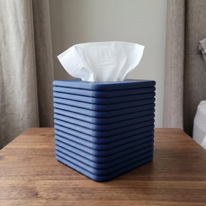 May include: A square, dark blue tissue box cover featuring a distinct, horizontally ribbed texture on all four sides. The cover is constructed with a flat top surface containing a central rectangular opening, through which white tissues are dispensed.