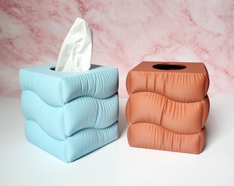 May include: Two decorative tissue box covers. One is light blue with a white tissue sticking out, and the other is a terracotta color. Both have a wavy, quilted design, and are square-shaped.