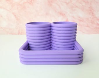 May include: A lavender-colored desk organizer with two cylindrical compartments and a rectangular base. The compartments are stacked with multiple circular layers. The base has a layered design, and the background is a pink and white marble pattern.