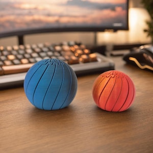 May include: Two spherical, 3D-printed objects sit on a wooden desk. One is blue, the other is orange. Both have a spiral groove design. The background includes a keyboard, computer monitor, and mouse.