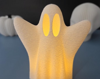 May include: A textured, off-white ghost-shaped lamp with glowing yellow eyes and a soft, warm light. The Halloween-themed decoration has outstretched arms and a friendly appearance. Other white decorative items are in the background.