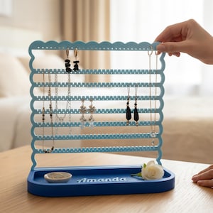 May include: A light blue jewelry organizer with a scalloped top and multiple horizontal bars for hanging earrings and necklaces. The base has a small dish and the name "Amanda" engraved. A white rose and jewelry are also present.