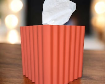 May include: A coral-colored tissue box with a ridged design. A white tissue is pulled from the top. The box is square and sits on a wooden surface. The background is blurred with soft, warm lighting.