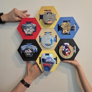 May include: A honeycomb-shaped medal display in various colors, holding several medals. The display is held by two sets of hands. The medals have different designs and ribbons, showcasing achievements from various events.