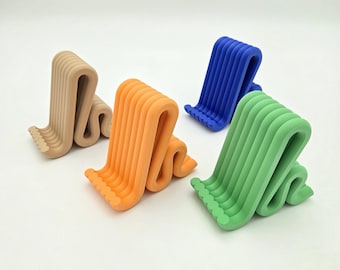 May include: Four wavy, S-shaped phone stands in beige, orange, blue, and light green. Each stand features a series of parallel, curved ridges that form a cradle for a mobile device, with a textured, scalloped edge along the base support area.