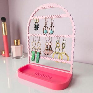 May include: A pink earring holder with a scalloped top and a tray base. The holder displays various earrings. The tray is embossed with the name "Natalie". A makeup brush and perfume bottle are visible in the background.