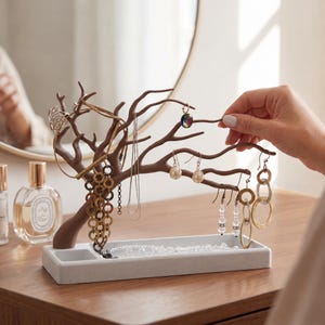 May include: A brown tree-shaped jewelry stand with multiple branches for hanging necklaces, earrings, and bracelets, featuring a rectangular light gray speckled base with a small recessed compartment for storing additional small accessories.