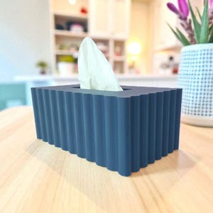 May include: A rectangular, navy blue tissue box with a ridged design. White tissues are visible, pulled from the top opening. The box sits on a light-colored wooden surface, with a blurred background of a kitchen.