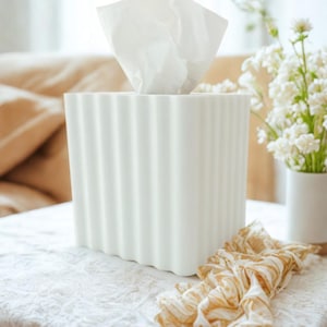 May include: A white tissue box with a ribbed design, holding white tissues. A patterned scarf in shades of gold and cream lies nearby. White flowers in a white vase are in the background. The scene is set on a white tablecloth.