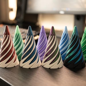 May include: A collection of eight cone-shaped decorative objects in various colors, including red, green, blue, and purple. Each cone features a spiral design with white accents, creating a visually appealing display. They are arranged on a dark wooden surface.