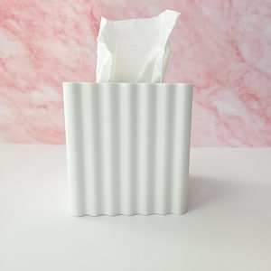 May include: A white tissue box with a ribbed design, holding a stack of white tissues. The background is a pink and white marble pattern. The tissue box is on a white surface.