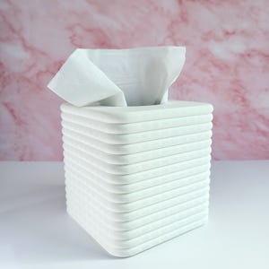 May include: White square tissue box holder with a white tissue box inside. The tissue box holder is made of stacked squares.