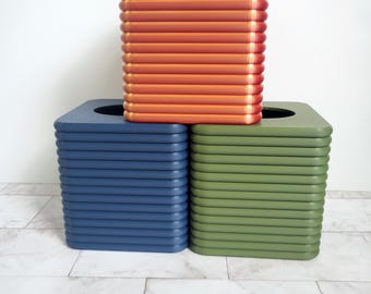 May include: Three square, stackable containers in shades of orange, blue, and green. The orange container sits atop the blue and green ones. Each container has a ridged, horizontal design. The containers are on a marble-patterned surface.